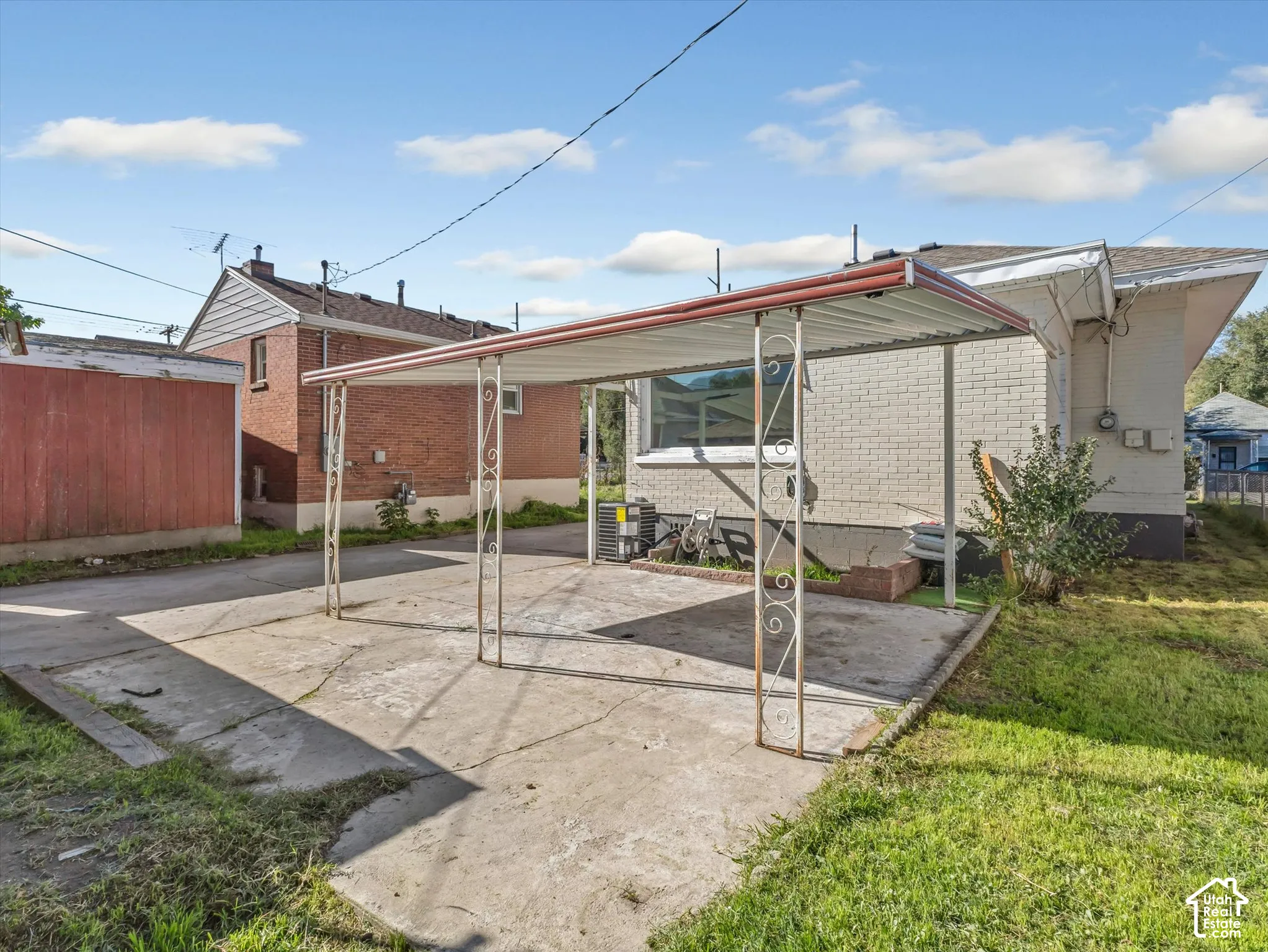 Exterior carport with metal roof structure and decorative wrought-iron supports, concrete driveway, and white brick facade