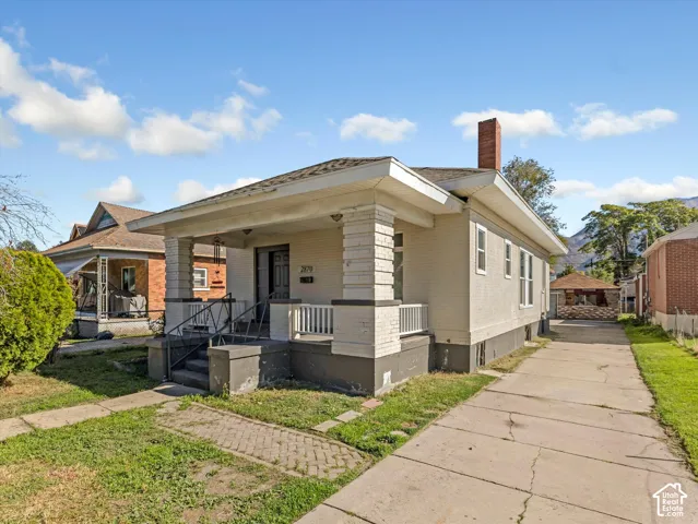 Single-story residence featuring light-colored masonry exterior, a covered front porch with tapered masonry pillars, a tall brick chimney, and a concrete driveway