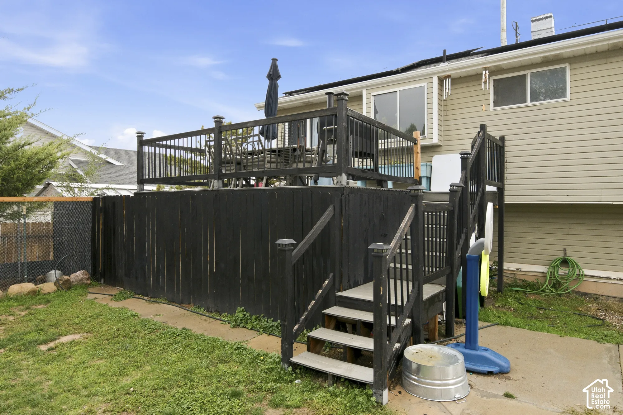 Raised wooden deck featuring black railing and stairs with access to a concrete patio and fenced backyard