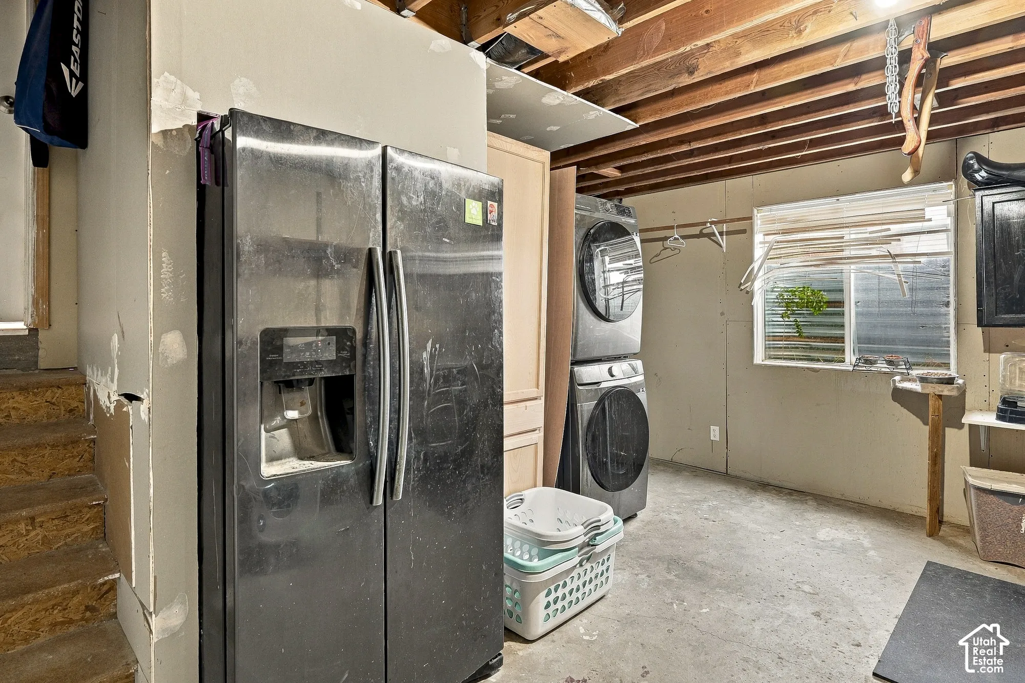 Laundry area featuring stacked metallic-finish washer and dryer, side-by-side refrigerator, concrete flooring, and exposed ceiling joists