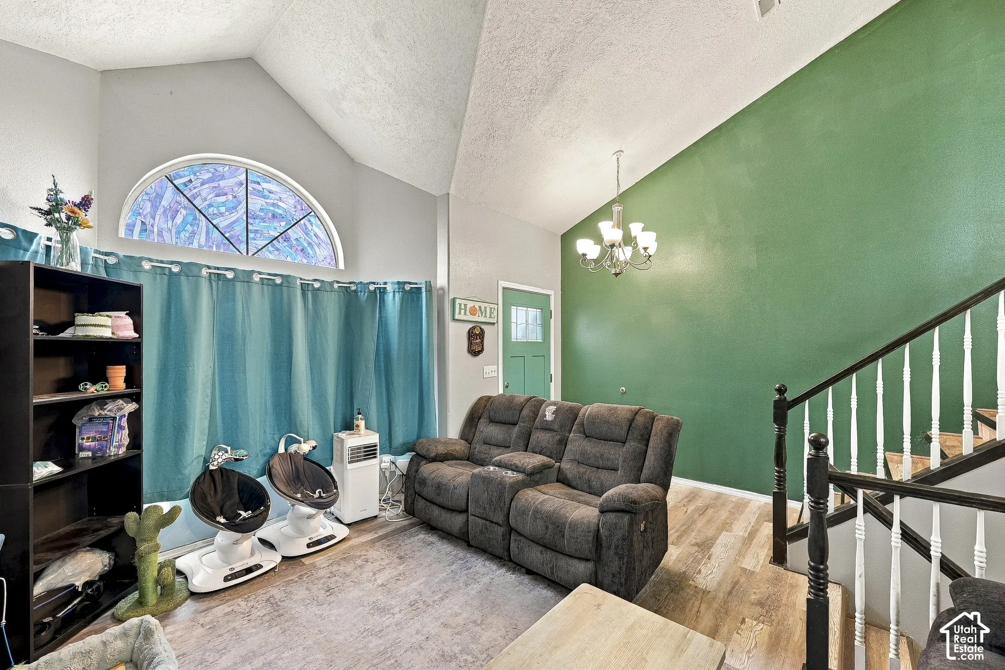 Vaulted ceilings, wood-finish flooring, an arched window, a multi-light chandelier, and a staircase featuring a white spindle railing with a black handrail and posts