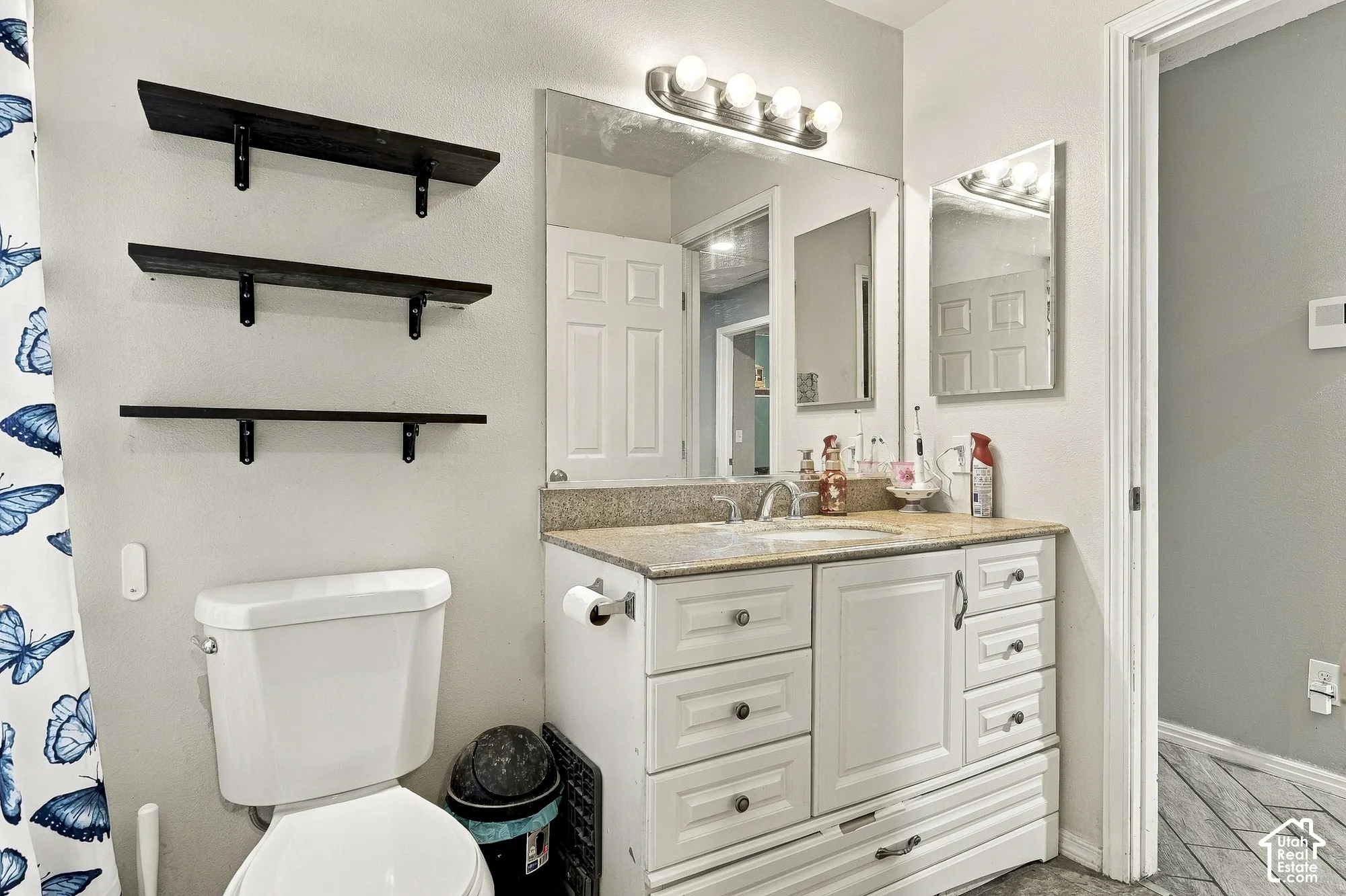 Bathroom vanity featuring white cabinetry, stone-finish countertop, undermount sink, four-light vanity fixture, and three dark-stained floating shelves