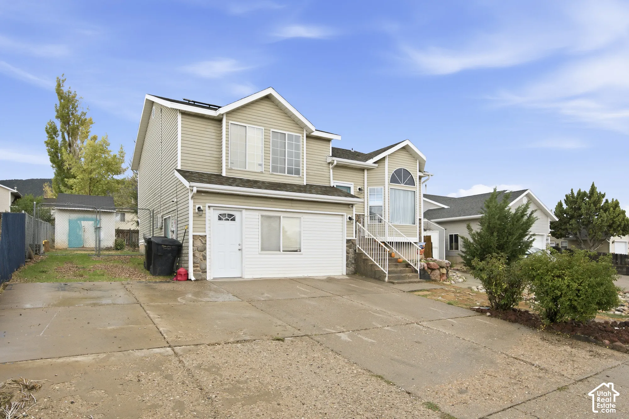 Two-story exterior featuring tan horizontal siding, stone veneer accents, an attached garage, a concrete driveway, and an elevated front staircase with metal railings