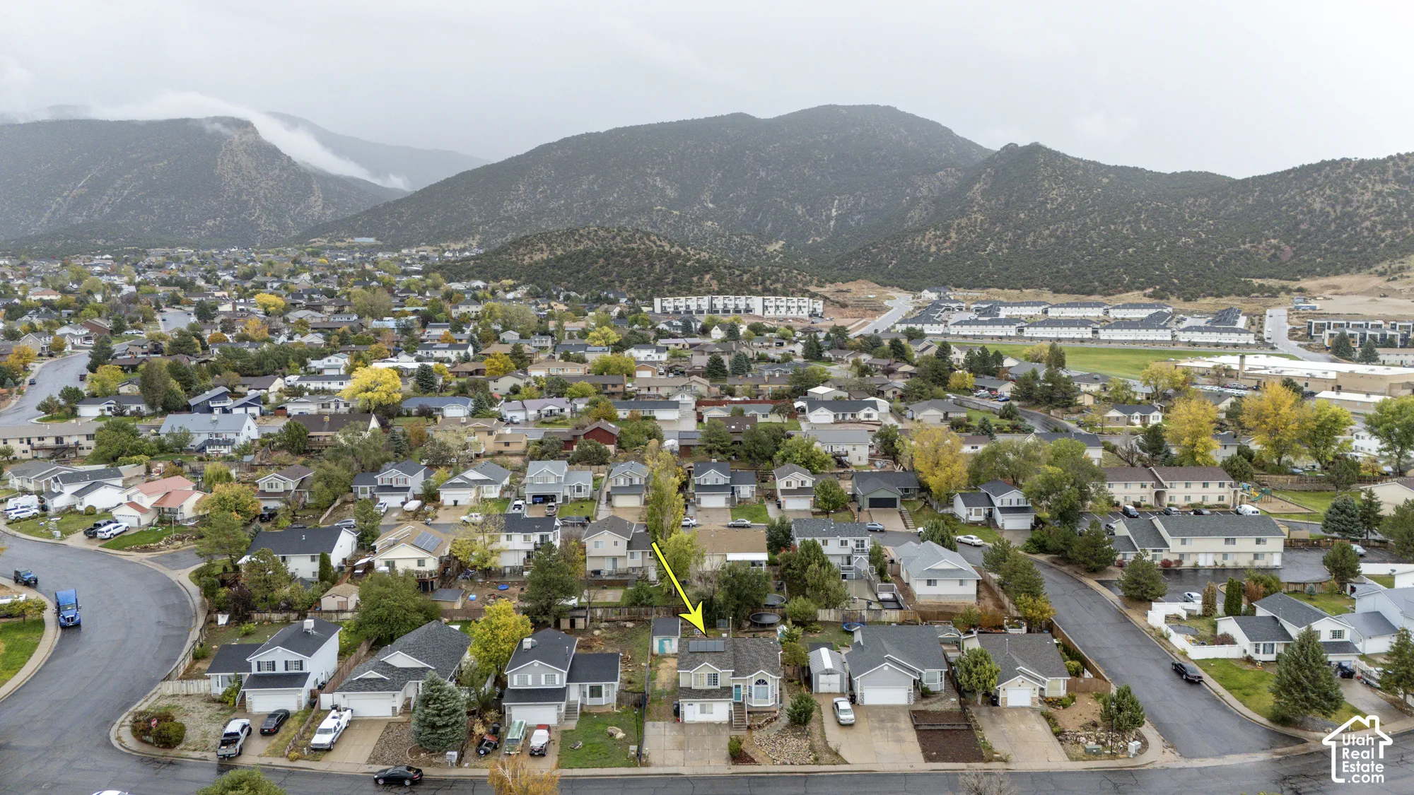 Aerial perspective of a detached residential property featuring a shingled roof, roof-mounted solar panels, an attached garage, and a backdrop of mountain scenery