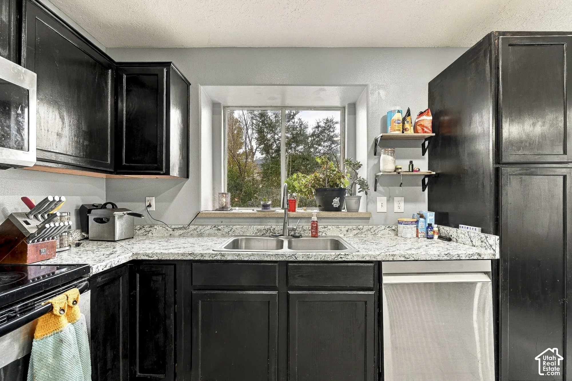 Kitchen featuring dark cabinetry, stone-surface countertops, a double-basin stainless steel sink, built-in dishwasher, and a recessed window