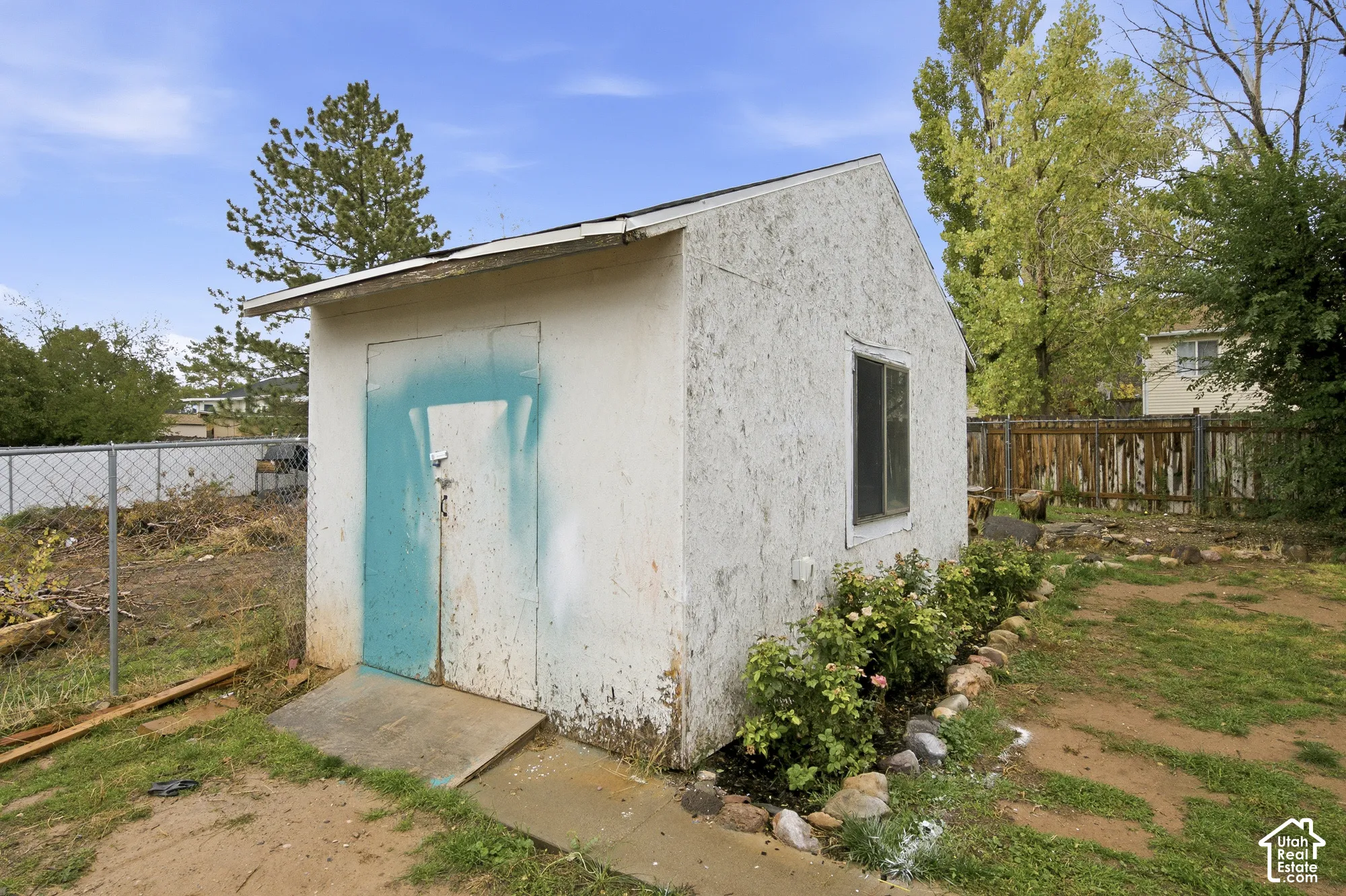 Detached structure featuring a textured exterior, gabled roof, and single-pane sliding window, set on a lot with perimeter fencing and natural landscaping