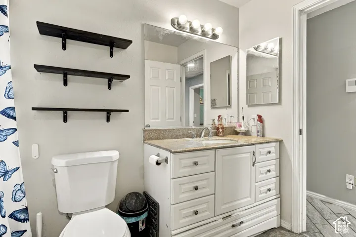 Bathroom vanity featuring white cabinetry, stone-finish countertop, undermount sink, four-light vanity fixture, and three dark-stained floating shelves