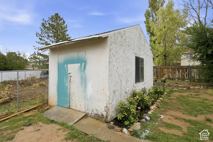 Detached structure featuring a textured exterior, gabled roof, and single-pane sliding window, set on a lot with perimeter fencing and natural landscaping