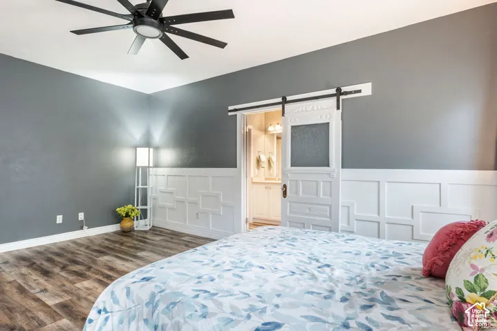 Room featuring wood-finish flooring, white wainscoting with geometric molding, a sliding barn-style door with frosted glass inlay, and a matte black ceiling fan