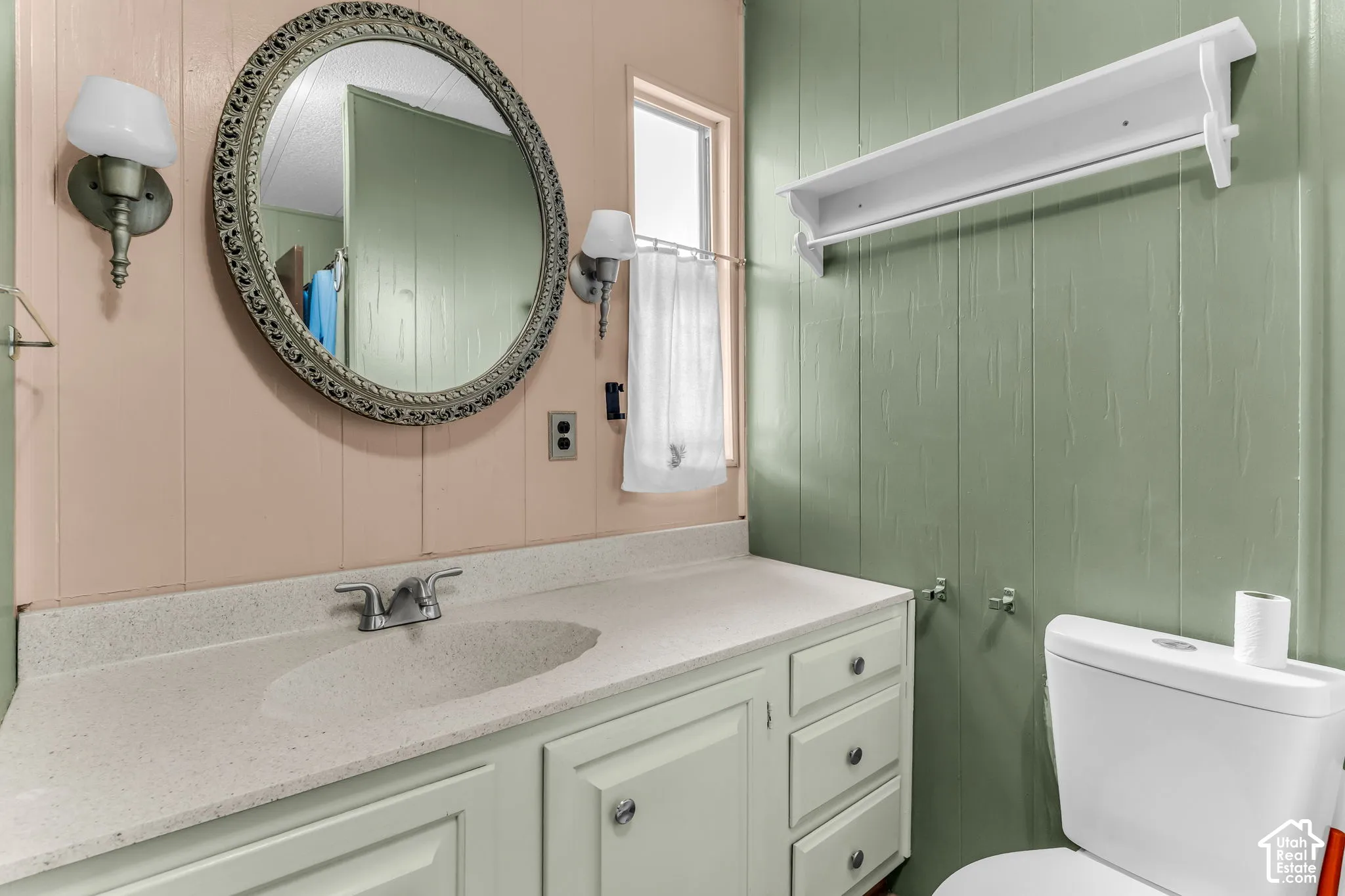 Bathroom vanity featuring a speckled countertop with an integrated basin, custom-painted cabinetry, decorative wall-mounted lighting, a circular framed mirror, and a fixed overhead shelf