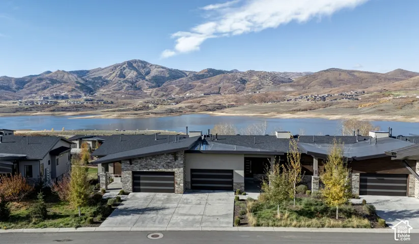 Contemporary multi-unit exterior featuring stacked stone masonry, horizontal metal panel accents, low-pitch shed rooflines, and concrete driveways, set against a backdrop of water and mountain terrain