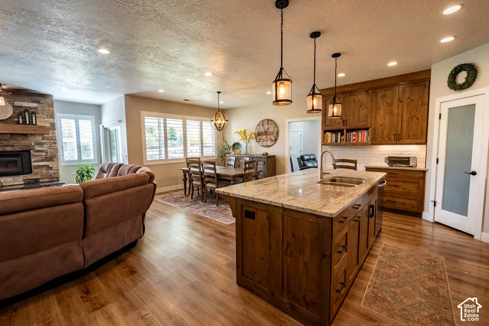 Kitchen featuring a stone-topped island with an integrated sink, natural wood cabinetry, pendant lighting, stone fireplace surround, and wood-finish flooring