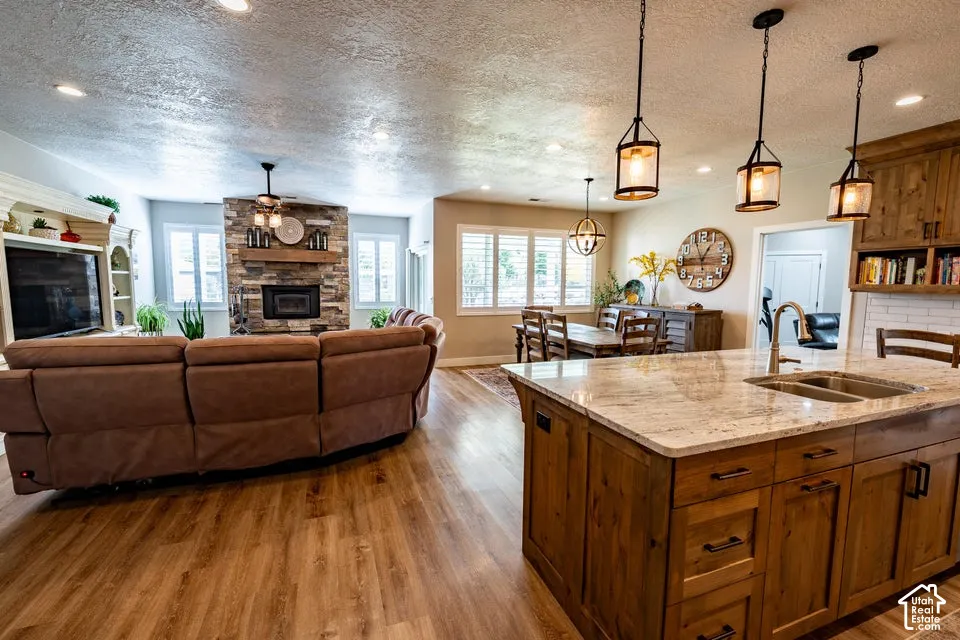 Kitchen island with stone countertop, under-mount stainless steel sink, wood-finish cabinetry, stone-clad fireplace with timber mantel, and wood-finish flooring