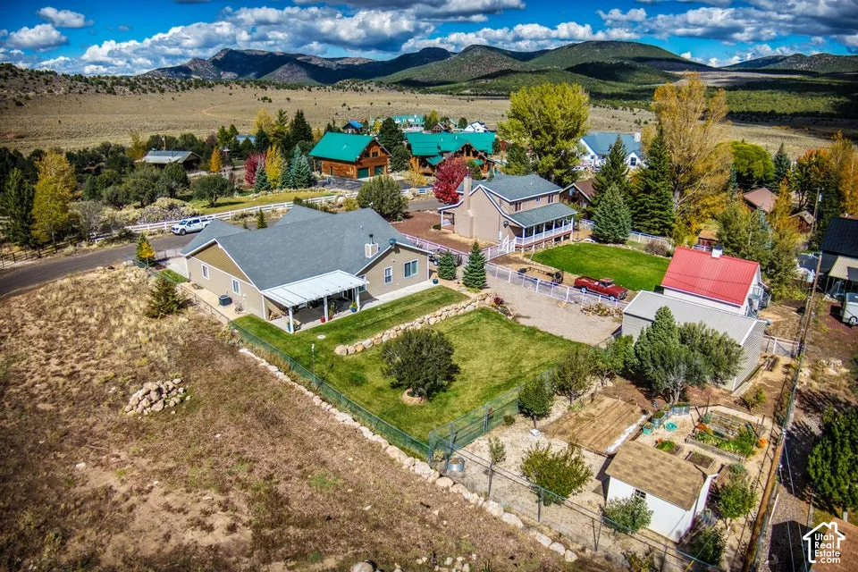 Aerial perspective of a property featuring a manicured lawn, fenced garden area, detached storage structure, covered patio, and mountain backdrop