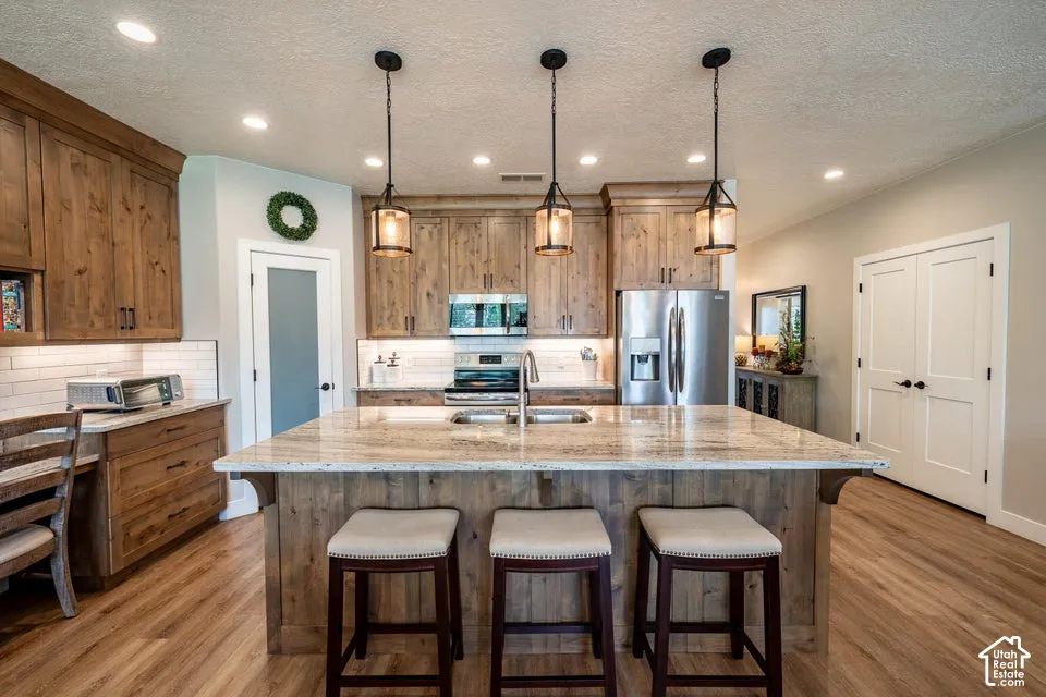 Kitchen featuring stained wood cabinetry, stone-surface central island with sink, white tile backsplash, stainless steel appliances, and wood-finish flooring