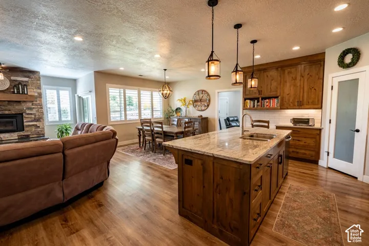 Kitchen featuring a stone-topped island with an integrated sink, natural wood cabinetry, pendant lighting, stone fireplace surround, and wood-finish flooring
