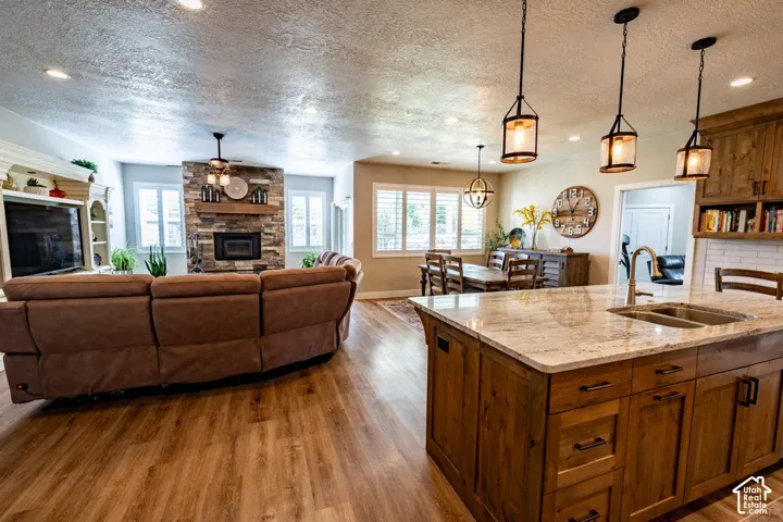 Kitchen island with stone countertop, under-mount stainless steel sink, wood-finish cabinetry, stone-clad fireplace with timber mantel, and wood-finish flooring