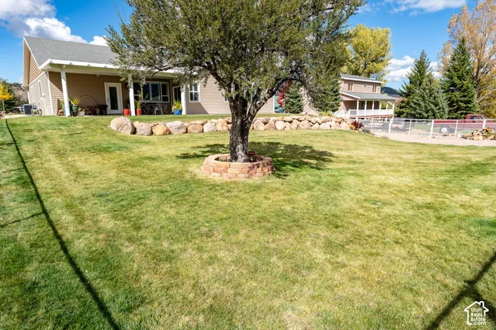 Expansive lawn featuring a mature tree with a circular stone planter, decorative boulder retaining wall, and covered patio area