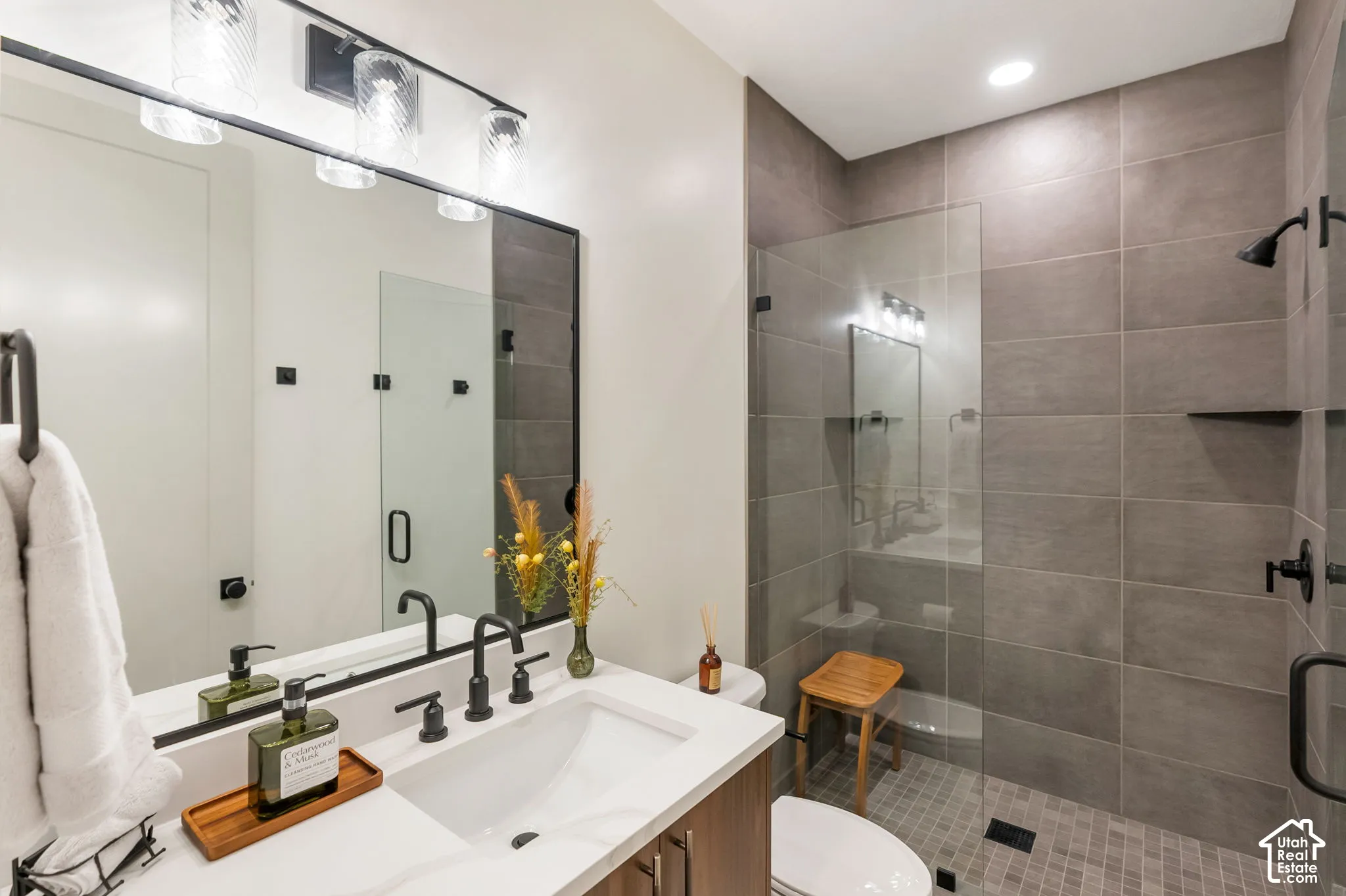 Bathroom featuring a wood-finish vanity with a white countertop, matte black fixtures, a framed wall mirror, and a walk-in shower with large-format wall tiles and a glass panel