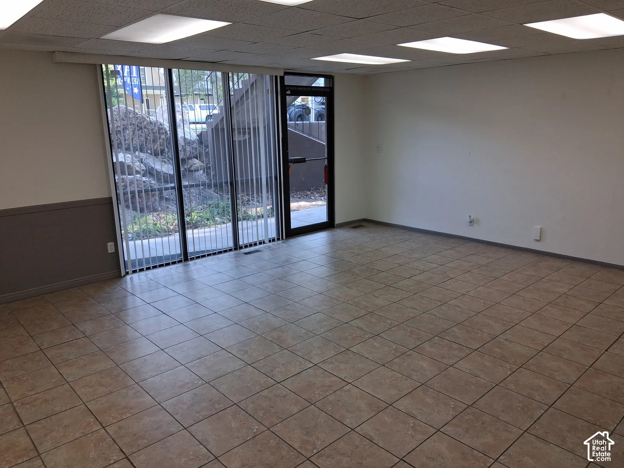Interior space featuring tile flooring, recessed panel ceiling, and large glass pane doors with vertical blinds