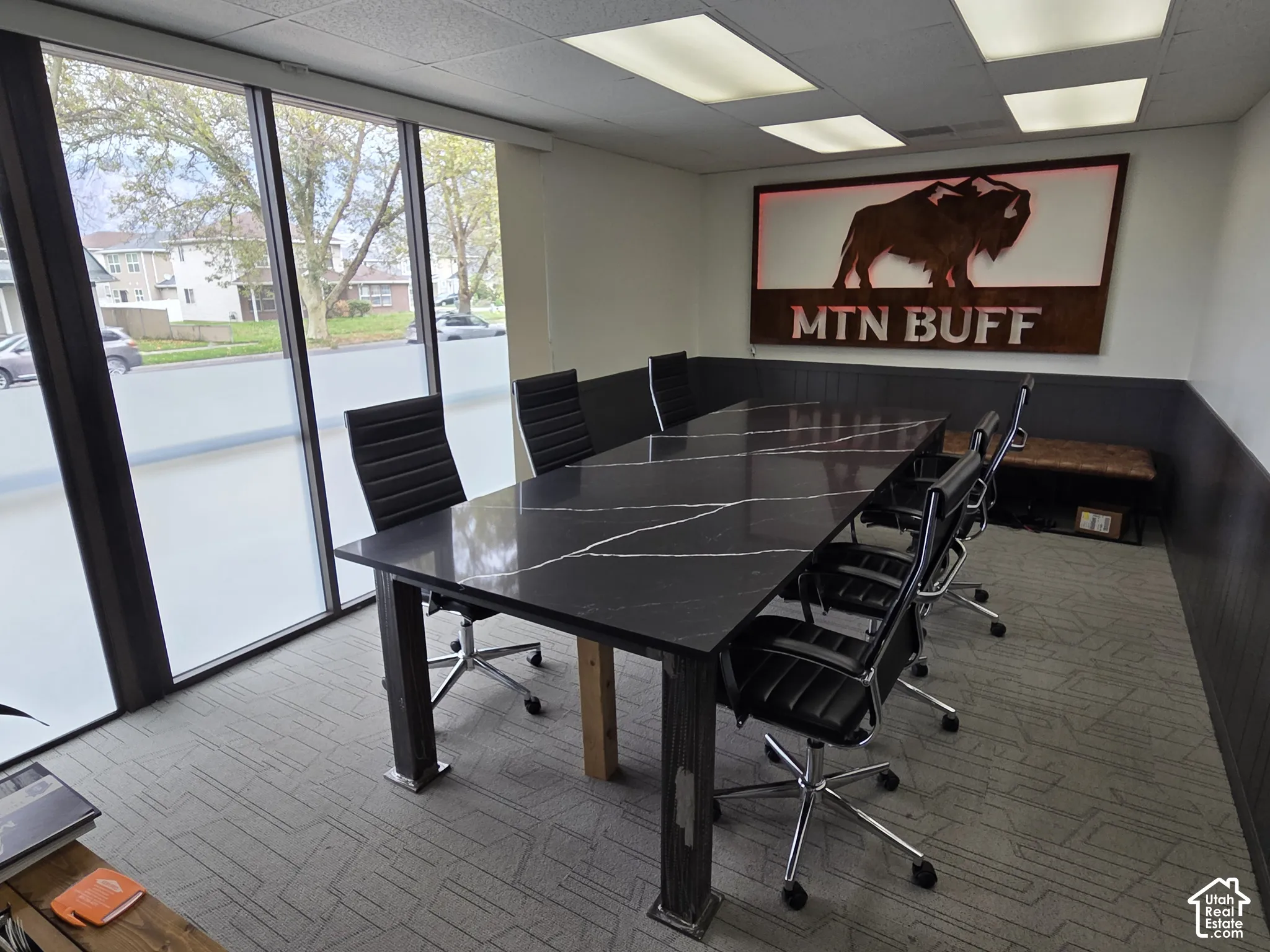 Conference room featuring floor-to-ceiling windows, dark-toned wall paneling, a large stone-surface table with industrial-style supports, and recessed panel lighting