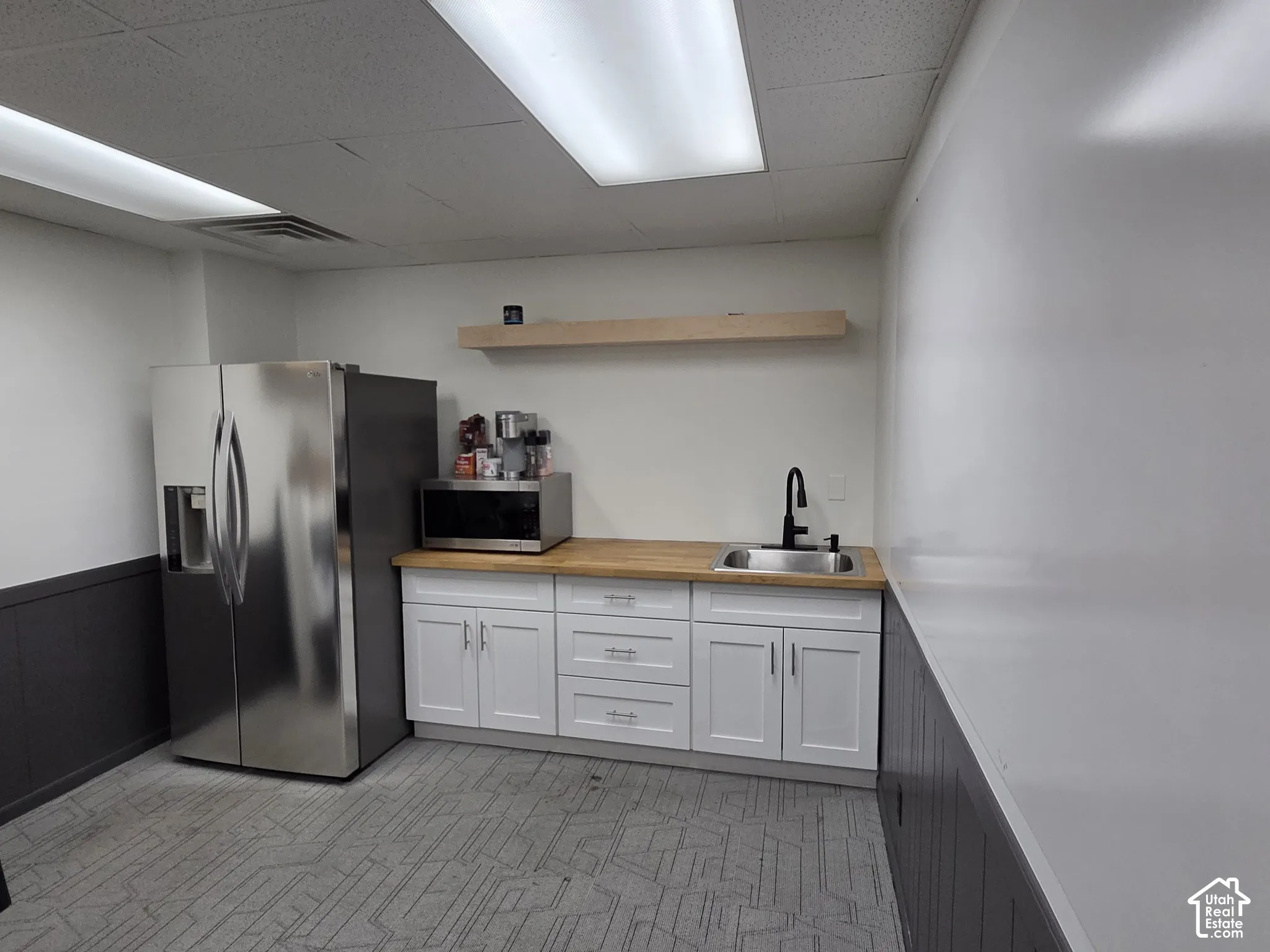 Stainless steel side-by-side refrigerator, white shaker-style cabinetry, wood-finish countertop, undermount stainless steel sink with matte black gooseneck faucet, and natural wood floating shelf