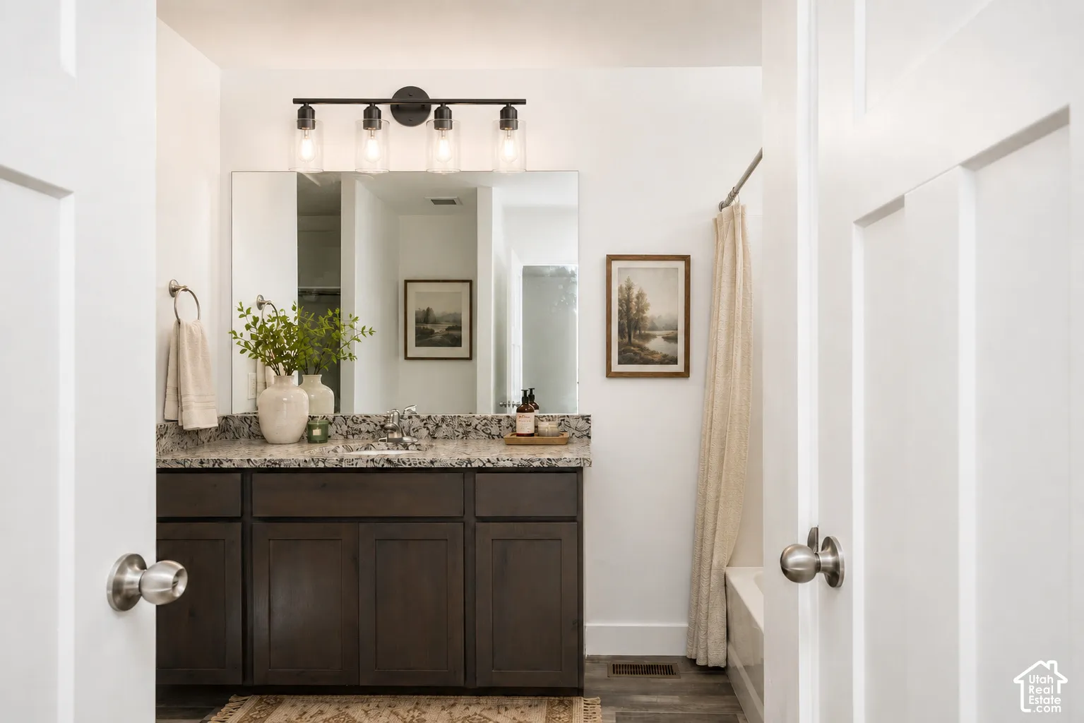 Bathroom featuring dark wood-finish cabinetry, a stone-pattern countertop with an integrated sink, a full-width vanity mirror, and a four-light fixture with glass shades