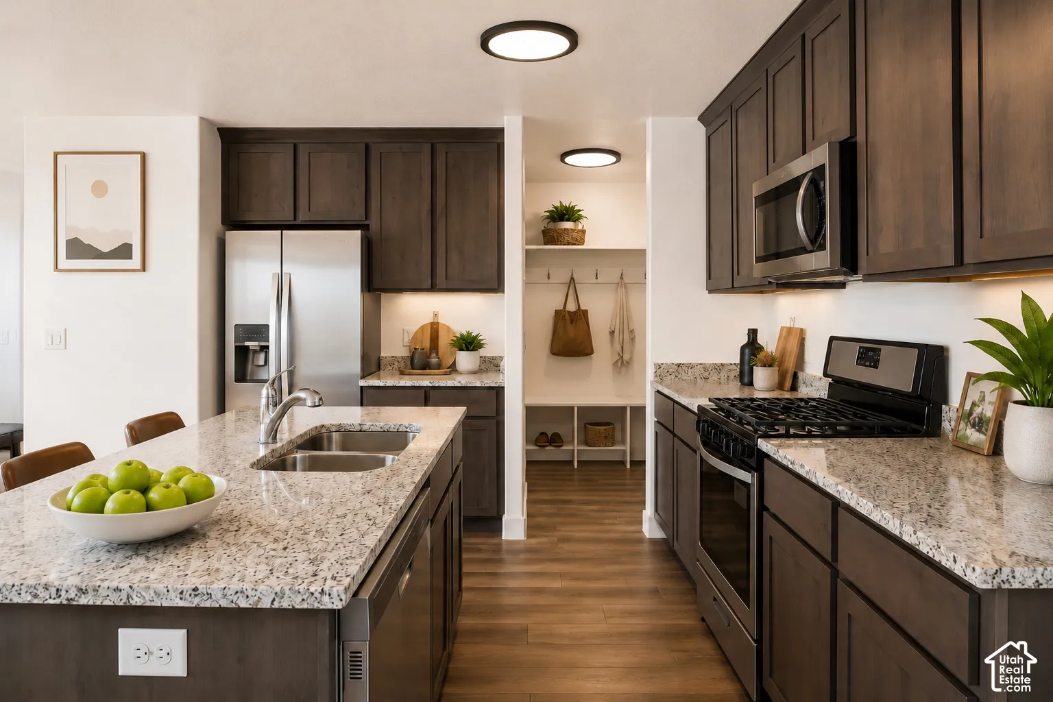 Kitchen featuring dark wood cabinetry, light-toned stone-finish countertops, stainless steel appliances, a center island with an undermount sink, and wood-finish flooring