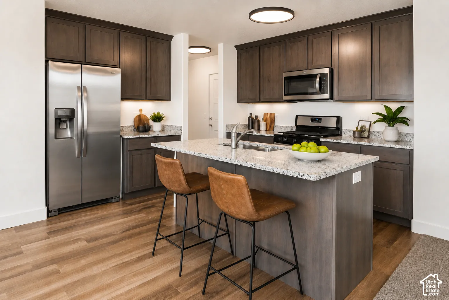 Kitchen featuring dark-toned cabinetry, light-colored stone-finish countertops, stainless steel appliances, a central island with an integrated sink, and wood-finish flooring