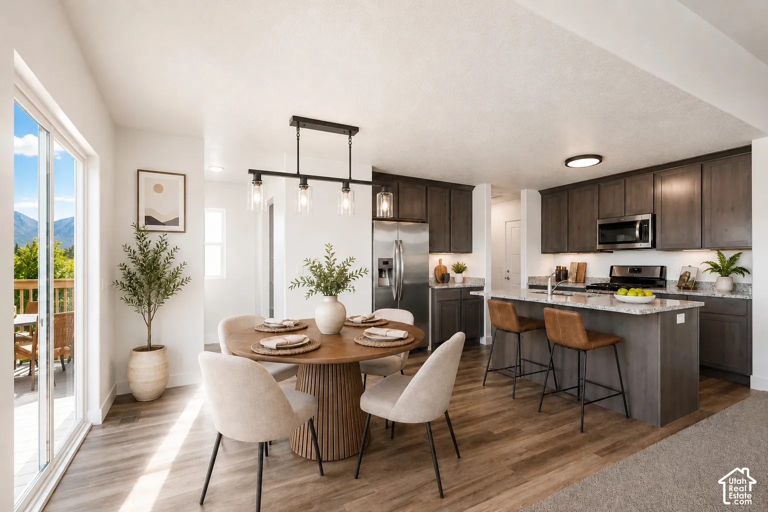 Open-concept kitchen featuring dark-stained shaker cabinetry, a stone-surfaced kitchen island with breakfast bar seating, stainless steel appliances, wood-finish flooring, and a multi-pendant light fixture