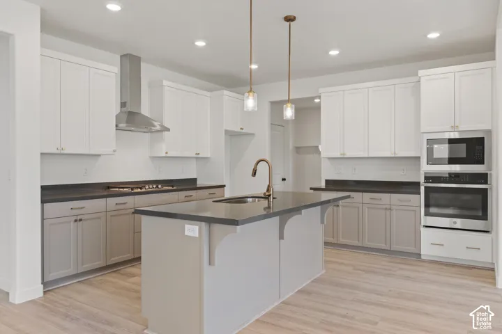 Kitchen featuring two-tone cabinetry, a central island with a prep sink, dark stone-surfaced countertops, a stainless steel chimney-style range hood, and wood-finish flooring
