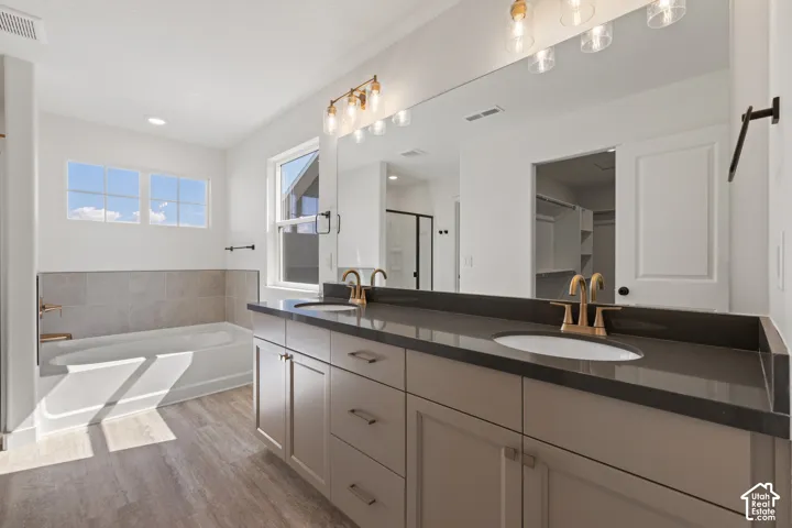 Dual-sink vanity with solid-surface countertop and metallic fixtures, recessed lighting, expansive wall-to-wall mirror, and a deep soaking tub featuring tiled surround and wood-finish flooring