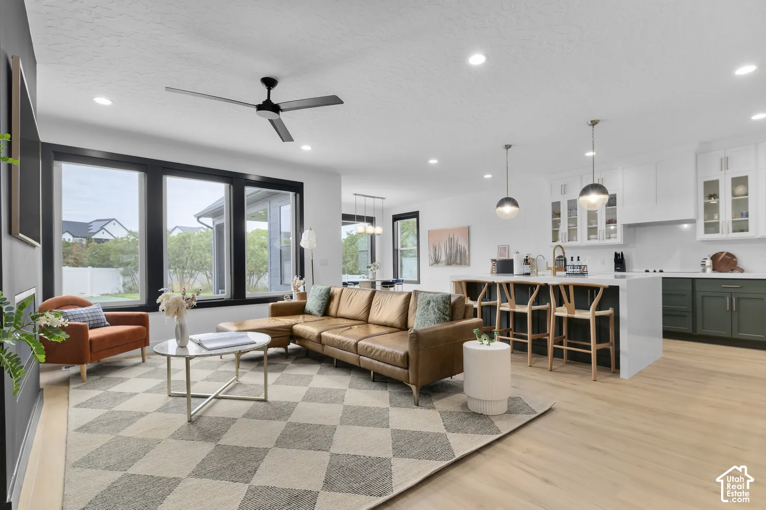 Open-concept interior featuring light wood-finish flooring, a kitchen island with waterfall-edge countertop, dark-toned lower cabinetry, large windows with black frames, and recessed lighting