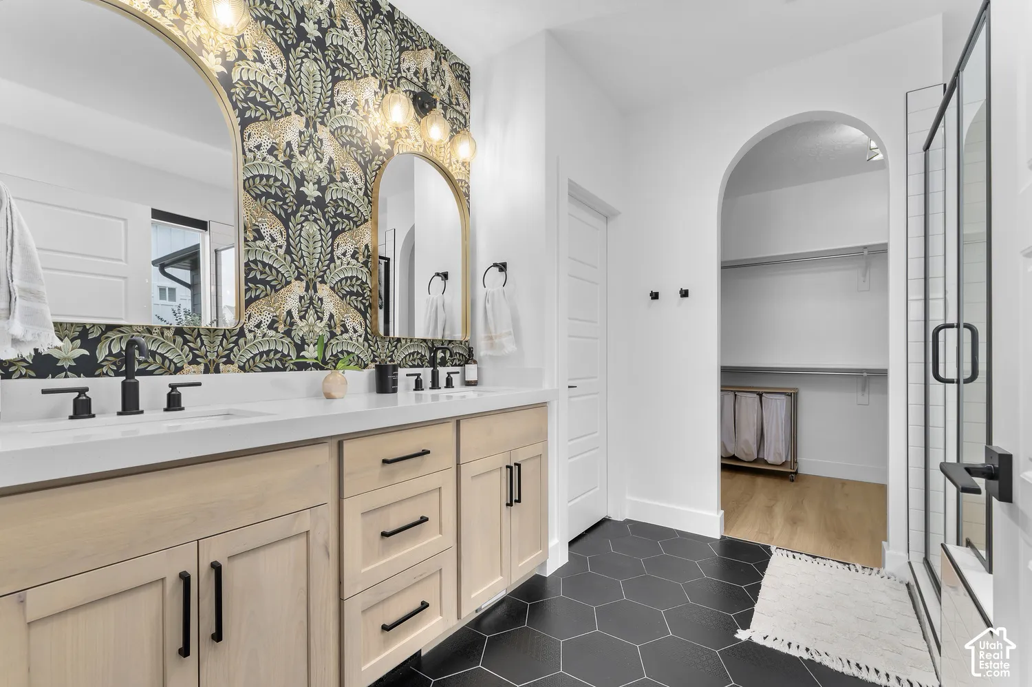 Dual-sink vanity with light-wood finish cabinetry, white countertop, matte black fixtures, arched vanity mirrors, and hexagonal floor tile