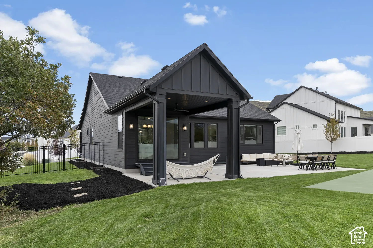 Exterior featuring a dark vertical-siding facade, a covered concrete patio, a built-in ceiling fan, black metal perimeter fencing, and a manicured lawn