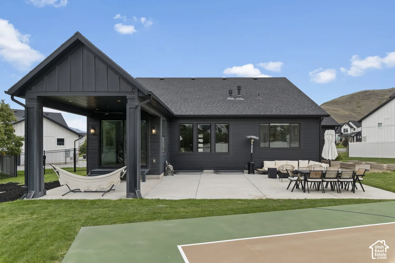 Rear exterior featuring dark board-and-batten siding, a covered patio with recessed lighting, a concrete patio area, a fenced-in yard, and a multi-sport court