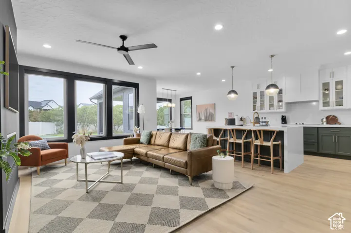 Open-concept interior featuring light wood-finish flooring, a kitchen island with waterfall-edge countertop, dark-toned lower cabinetry, large windows with black frames, and recessed lighting
