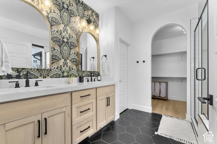 Dual-sink vanity with light-wood finish cabinetry, white countertop, matte black fixtures, arched vanity mirrors, and hexagonal floor tile