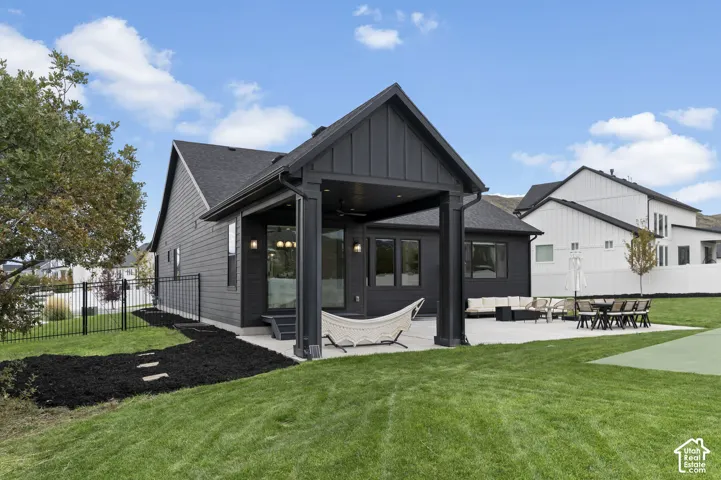 Exterior featuring a dark vertical-siding facade, a covered concrete patio, a built-in ceiling fan, black metal perimeter fencing, and a manicured lawn