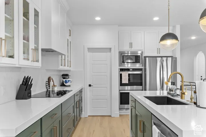 Kitchen featuring white shaker-style cabinetry with glass-front uppers, solid surface countertops, a center island with an undermount sink and metallic finish faucet, stainless steel built-in appliances, and wood-finish flooring