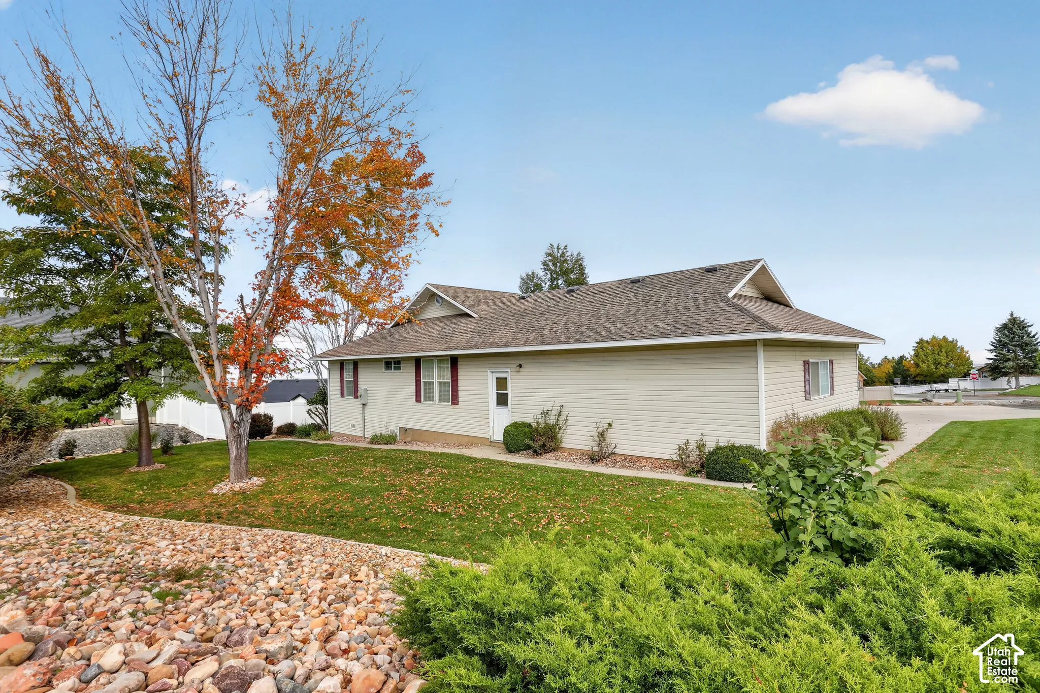 Exterior featuring light-colored horizontal siding, asphalt shingle roofing, mature landscape trees, and rock-mulched ground cover