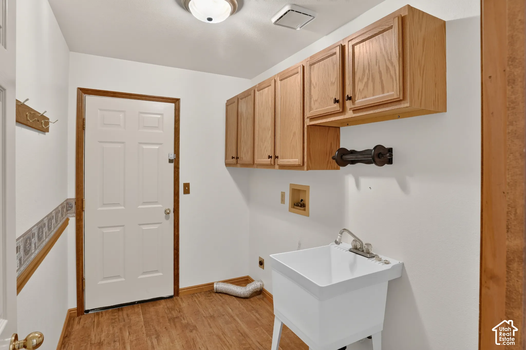 Laundry space featuring wood-finish flooring, white utility sink with chrome faucet, upper wood cabinetry, and a wall-mounted wood hook rack