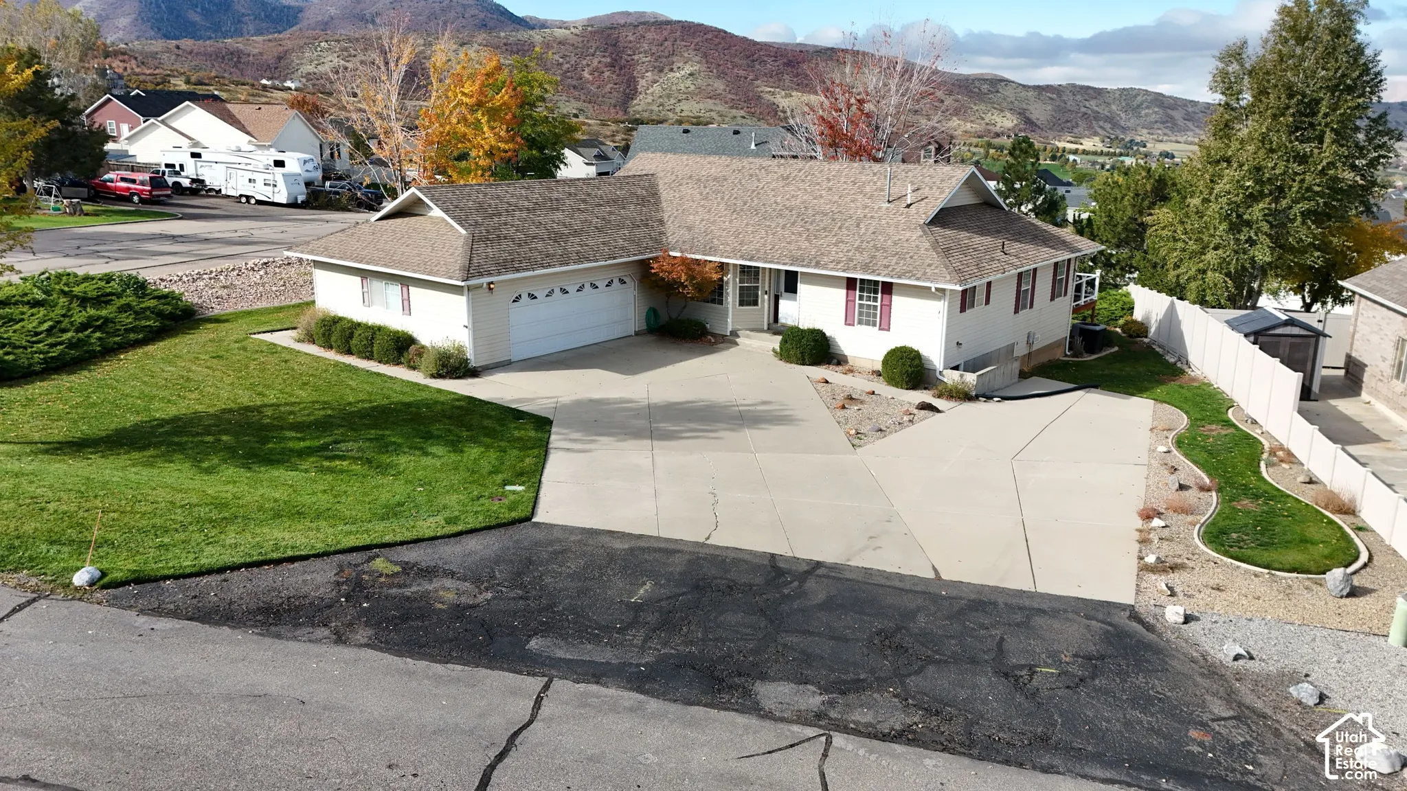 Single-story residence featuring light-colored siding, an attached two-car garage, an expansive concrete driveway, and a landscaped front lawn with mountain surroundings