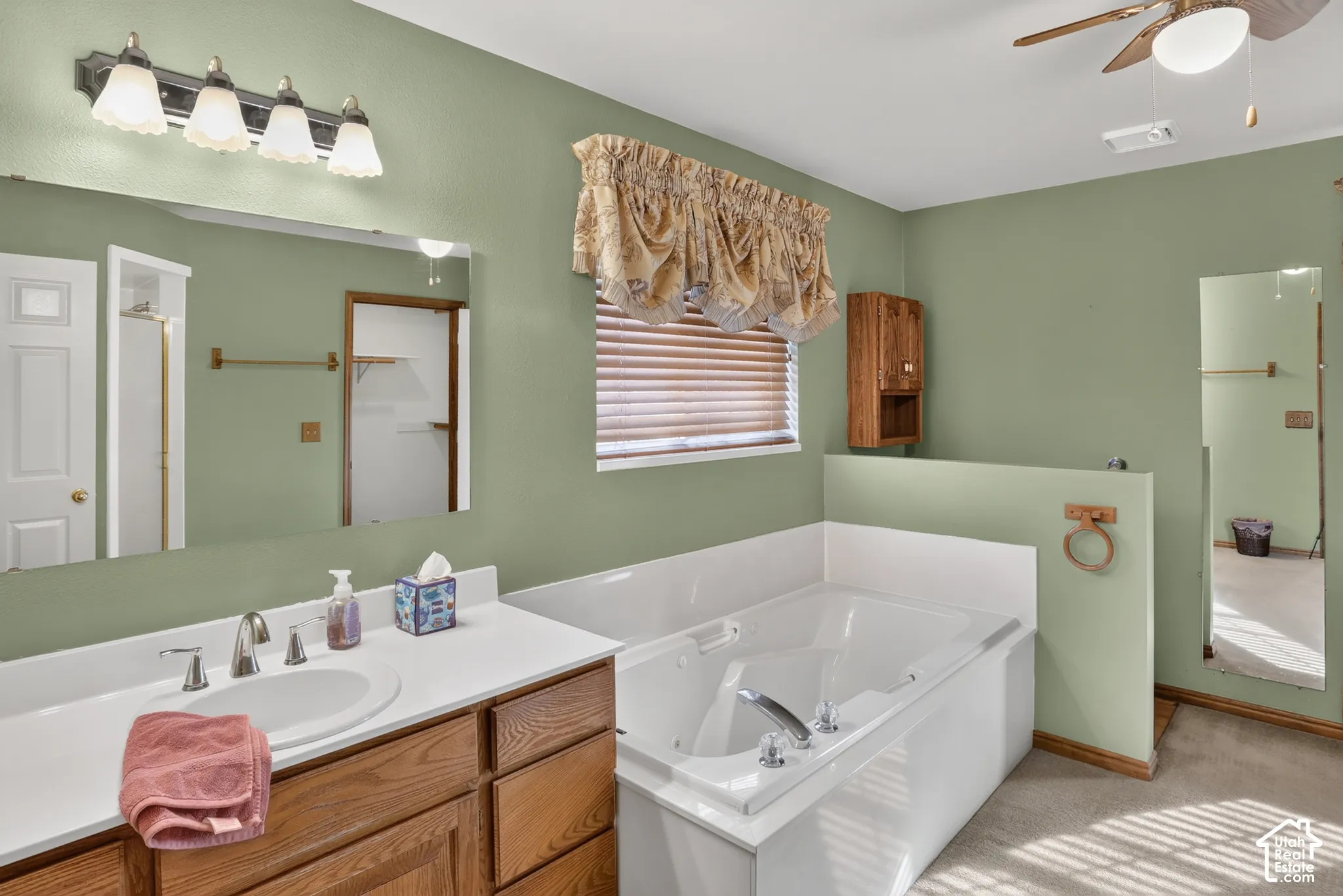 Bathroom featuring a wooden vanity with a white countertop, inset sink, large wall-mounted mirror, soaking tub with tile surround, and a ceiling-mounted fan with light fixture