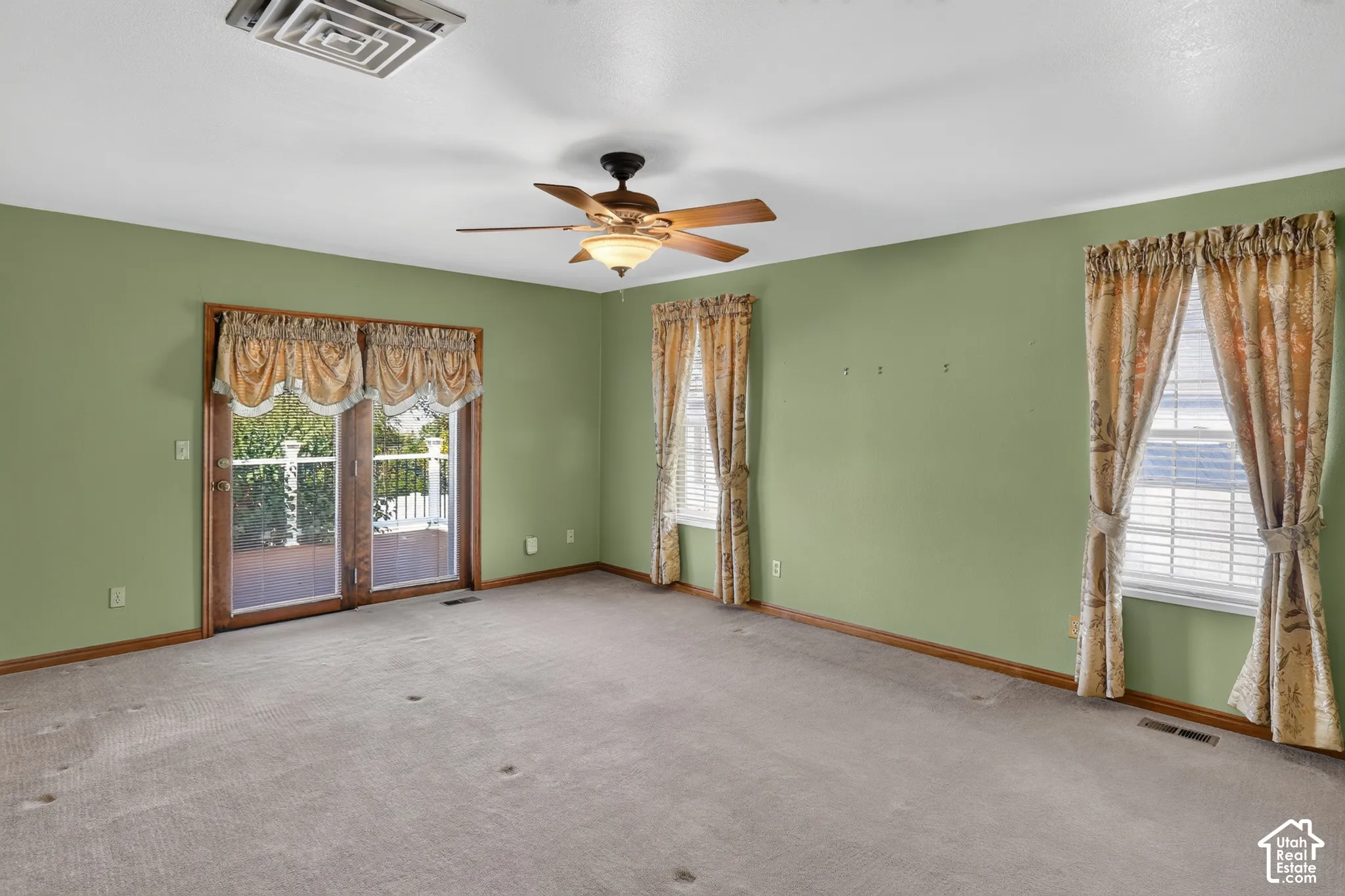 Spacious room featuring neutral carpeting, a centrally mounted ceiling fan with integrated lighting, dual-pane windows, and wood-framed sliding glass doors