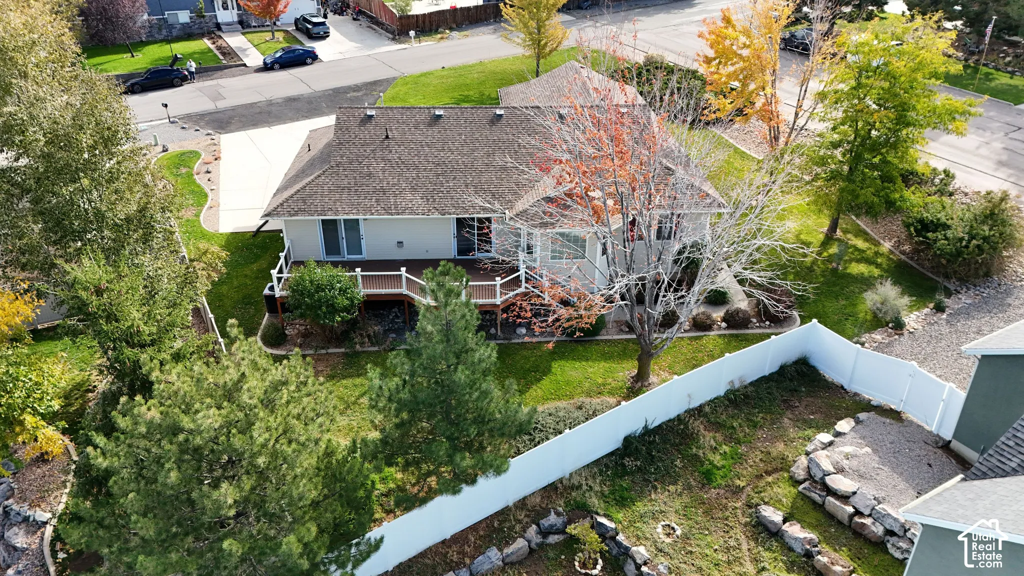 Elevated view of a residential property featuring an expansive elevated wooden deck with railing, a fenced backyard, mature trees, and a concrete driveway