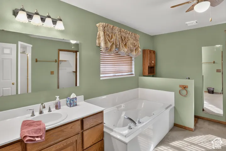 Bathroom featuring a wooden vanity with a white countertop, inset sink, large wall-mounted mirror, soaking tub with tile surround, and a ceiling-mounted fan with light fixture
