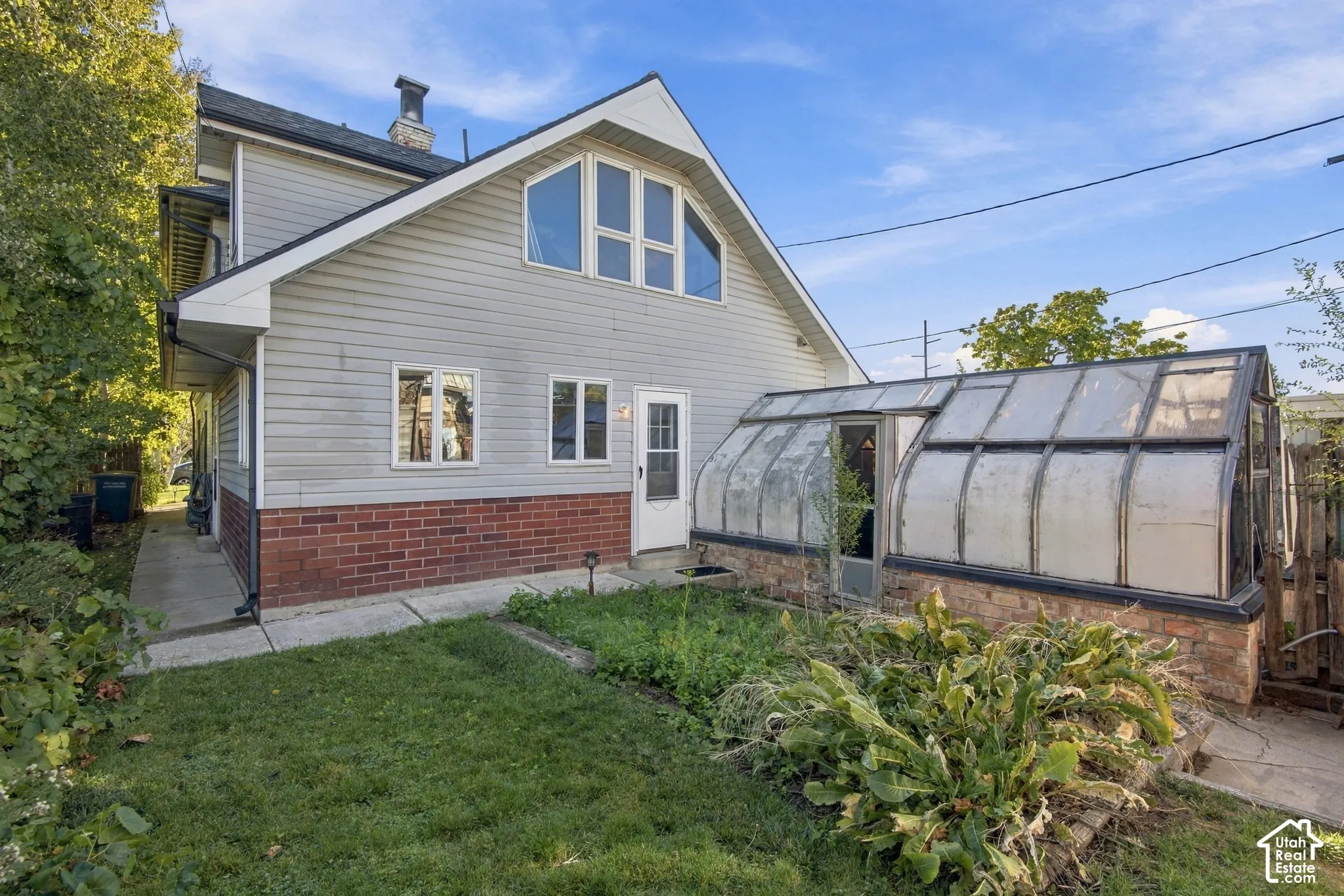 Exterior featuring horizontal siding, a red brick base, an integrated glass greenhouse, multiple rectangular windows, and a concrete pathway