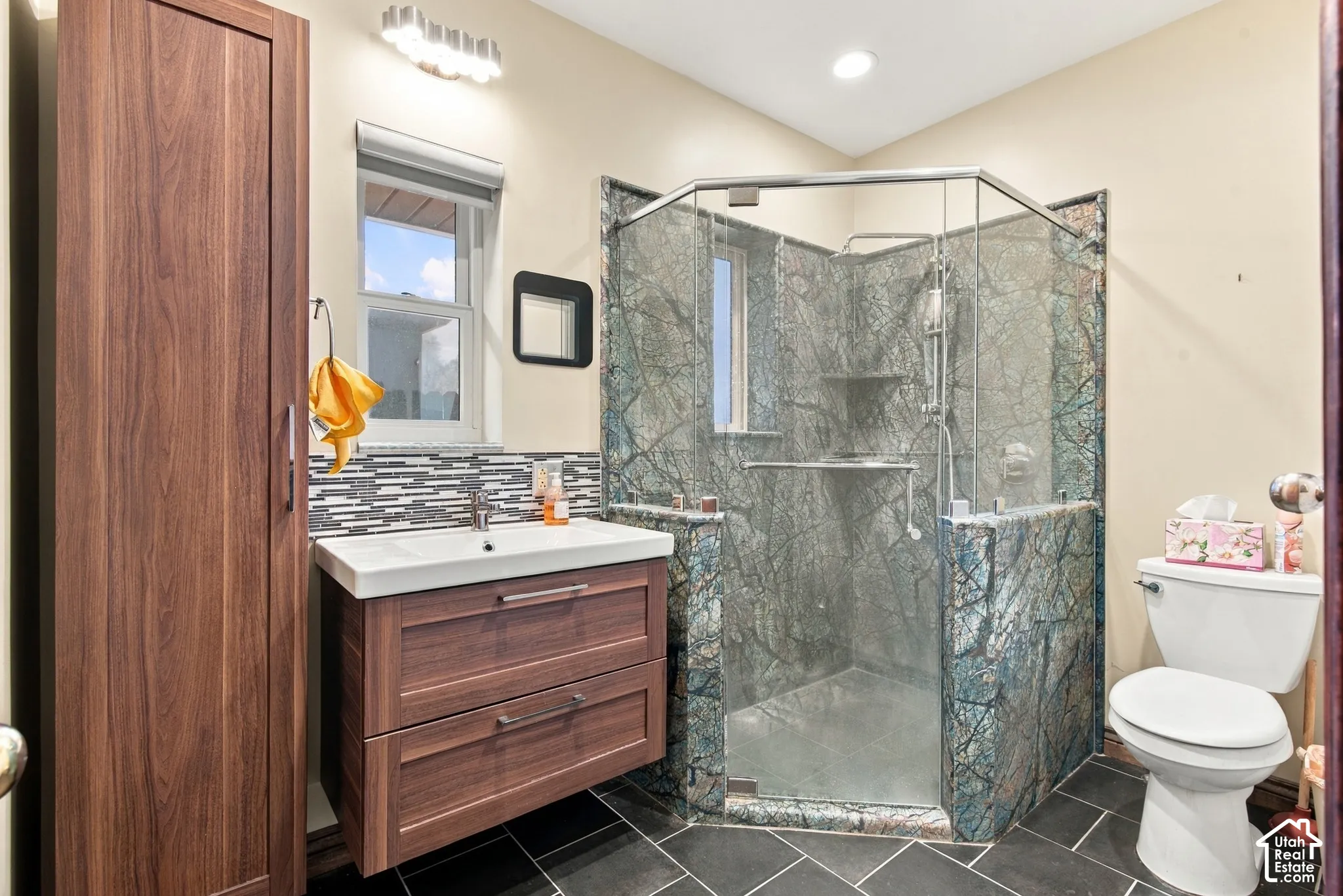 Bathroom featuring a floating wood-finish vanity, a corner glass-enclosed shower with decorative stone-patterned tile surround, a mosaic tile backsplash, and dark tile flooring