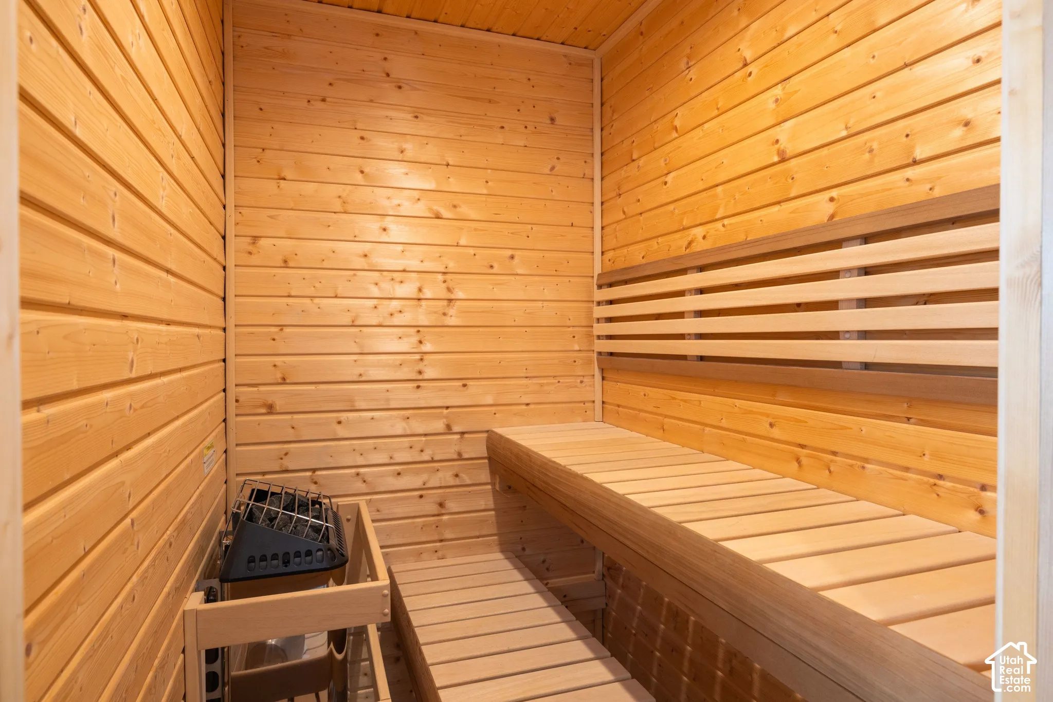 Sauna featuring natural wood tongue-and-groove wall paneling, built-in tiered wooden benches, and an integrated stone-filled electric heater with a protective wooden guard